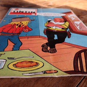 May include: A comic book cover featuring a scene with two cartoon characters in a room. One character is sweating profusely, while the other stands with arms crossed. The word "TINTIN" is at the top. A table is set with food.