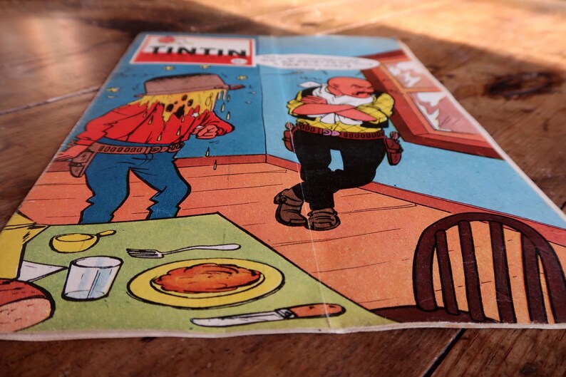 May include: A comic book cover featuring a scene with two cartoon characters in a room. One character is sweating profusely, while the other stands with arms crossed. The word "TINTIN" is at the top. A table is set with food.
