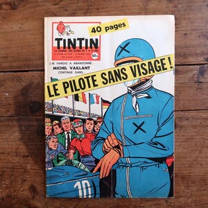 May include: Vintage Tintin comic book cover featuring a race car driver in a blue jumpsuit and helmet. The cover has the title "LE PILOTE SANS VISAGE!" and the Tintin logo. The comic is on a wooden surface.