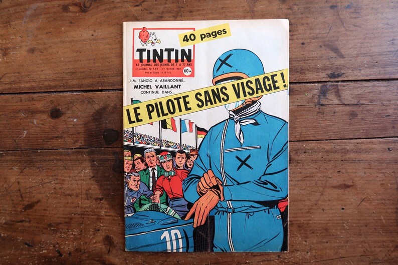 May include: Vintage Tintin comic book cover featuring a race car driver in a blue jumpsuit and helmet. The cover has the title "LE PILOTE SANS VISAGE!" and the Tintin logo. The comic is on a wooden surface.