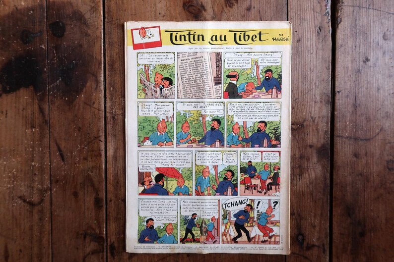 May include: A vintage comic book page titled "Tintin au Tibet" featuring a series of illustrated panels. The panels depict scenes with characters in various settings, with speech bubbles containing text. The comic is set against a wooden background.