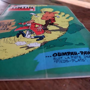 May include: A vintage Tintin comic book titled Sur le sentier de la guerre featuring a green cover with a large yellow footprint illustration. The artwork depicts a character in a red coat and a figure in brown clothing with the text Oumpah-pah.