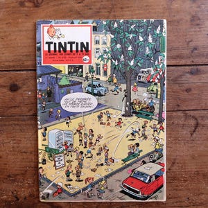 Puede incluir: Una portada de c&oacute;mic vintage que representa una bulliciosa escena callejera con numerosas figuras, coches y edificios. El t&iacute;tulo "TINTIN" aparece en la parte superior, con texto en franc&eacute;s debajo. La portada est&aacute; sobre un fondo de madera.