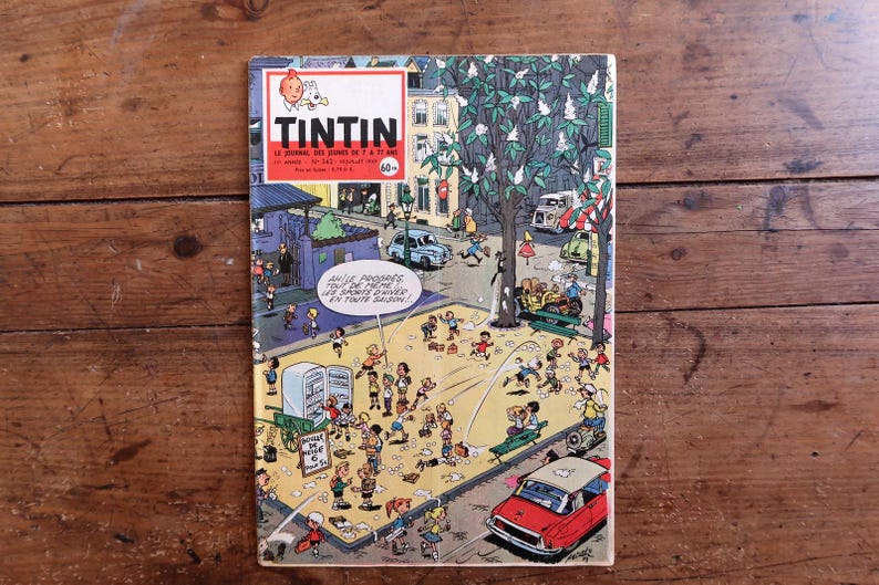 Puede incluir: Una portada de c&oacute;mic vintage que representa una bulliciosa escena callejera con numerosas figuras, coches y edificios. El t&iacute;tulo "TINTIN" aparece en la parte superior, con texto en franc&eacute;s debajo. La portada est&aacute; sobre un fondo de madera.