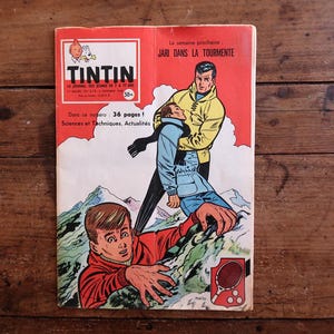 May include: A vintage Tintin comic book cover, depicting a dramatic scene with two men on a mountain. The cover is red and white, with the title "TINTIN" at the top. The comic book is resting on a wooden surface.
