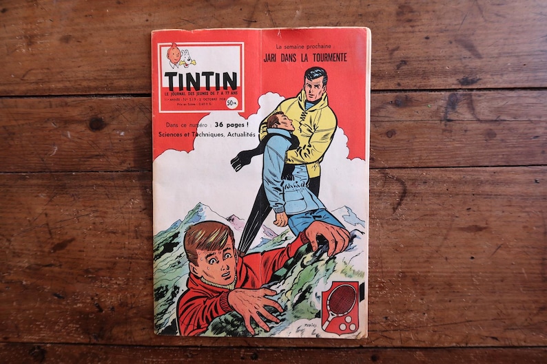 May include: A vintage Tintin comic book cover, depicting a dramatic scene with two men on a mountain. The cover is red and white, with the title "TINTIN" at the top. The comic book is resting on a wooden surface.