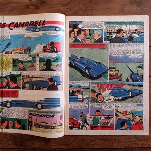 May include: Open book with vintage comic strip illustrations. The comic features scenes with blue race cars, people, and text bubbles. The title "LES CAMPBELL" is visible at the top. The pages are on a wooden surface.