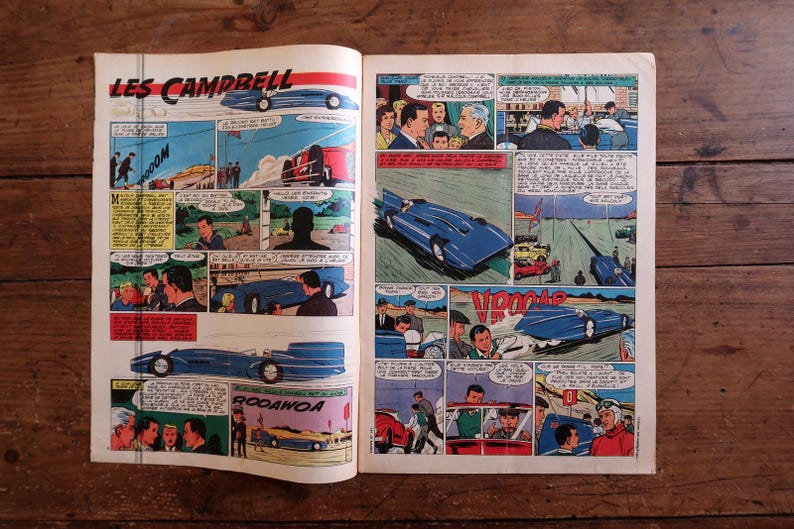 May include: Open book with vintage comic strip illustrations. The comic features scenes with blue race cars, people, and text bubbles. The title "LES CAMPBELL" is visible at the top. The pages are on a wooden surface.