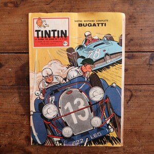 May include: A vintage Tintin comic magazine with a yellow cover featuring a blue Bugatti race car numbered 13 in the foreground and a light blue race car numbered 28 in the background. The text reads Tintin, Notre Histoire Complete: Bugatti.
