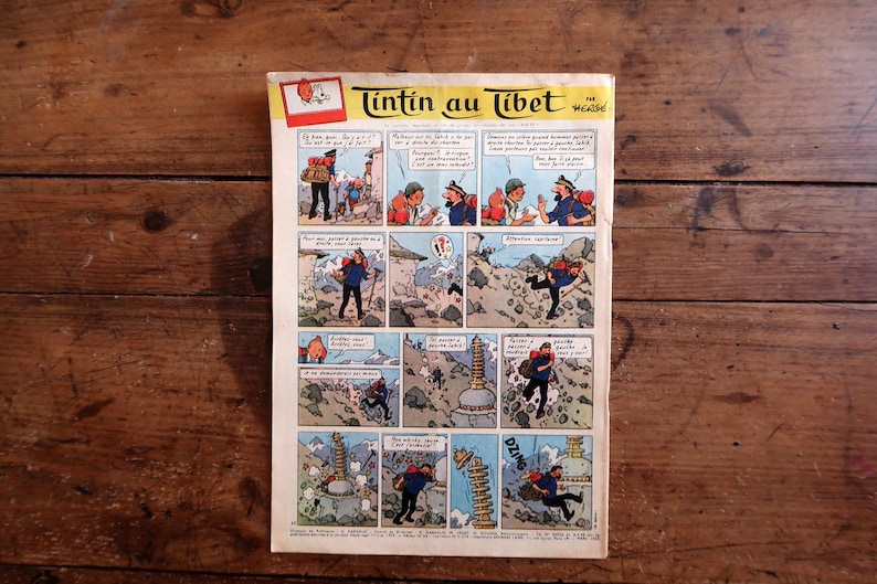 May include: A vintage comic book page titled Tintin au Tibet featuring a multi-panel color illustration. The comic strip depicts characters in a mountainous landscape with dialogue bubbles in French. The paper has a cream background with aged edges.