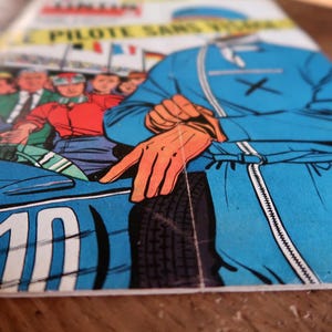 May include: Comic book cover featuring a racing theme. The illustration depicts a blue race car with the number 10, a driver in a blue racing suit, and a crowd of spectators. The comic book title is partially visible.