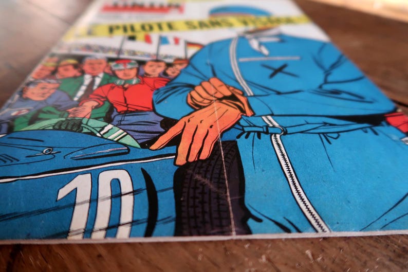 May include: Comic book cover featuring a racing theme. The illustration depicts a blue race car with the number 10, a driver in a blue racing suit, and a crowd of spectators. The comic book title is partially visible.