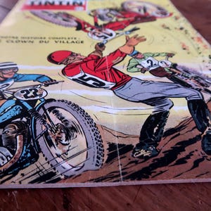 May include: Comic book cover featuring motorcycle racers in action. The illustration shows racers in red, blue, and green outfits, with the number 23 visible on a motorcycle. The title "LE CLOWN DU VILLAGE" is also visible.