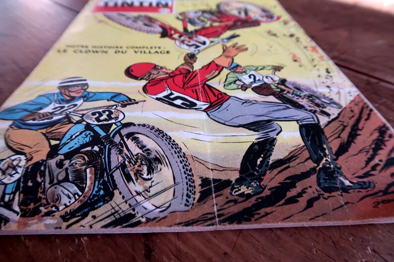 May include: Comic book cover featuring motorcycle racers in action. The illustration shows racers in red, blue, and green outfits, with the number 23 visible on a motorcycle. The title "LE CLOWN DU VILLAGE" is also visible.