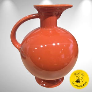 May include: A vibrant orange ceramic pitcher with a rounded body, a curved handle, and a spout. The pitcher has a glossy finish and sits on a small, circular base. The overall design is simple and elegant, suitable for serving beverages.