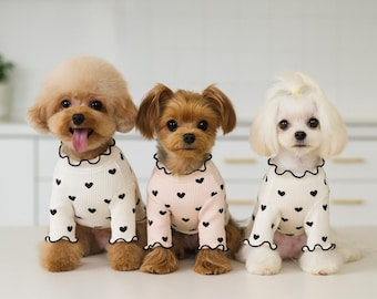 May include: Three small dogs wearing matching cream-colored sweaters with black heart patterns. Each sweater has a ruffled collar and cuffs. The dogs are sitting on a white surface, with a white background.