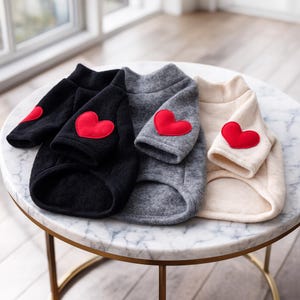 May include: Four dog sweaters in black, charcoal gray, and cream colors, each with a red heart applique on the sleeve. The sweaters are made of a soft, textured fabric and are displayed on a marble-topped table.
