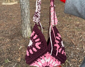 May include: A handmade crochet bag in shades of burgundy, pink, and white. The bag features a geometric pattern and a matching handle. The bag is held by a person in a gray coat with a red sleeve.
