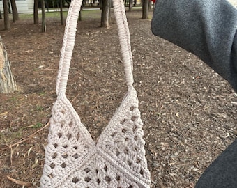 May include: A beige crochet shoulder bag with a geometric openwork design. The bag features a triangular shape and long, braided handles. The bag is held up by a person wearing a gray coat with a red sweater.