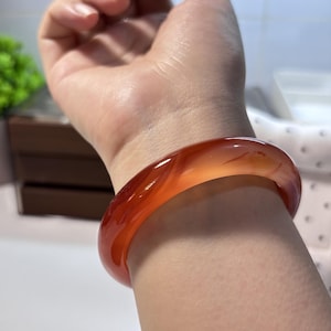 Op de afbeelding: Een gladde, doorschijnende oranje-rode carneool armband met een gepolijste afwerking. De cirkelvormige band vertoont natuurlijke kleurvariaties en interne insluitingen, wat zorgt voor een levendige, warme tint die het licht opvangt.