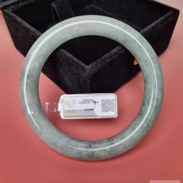 May include: A smooth, circular jadeite bangle with a pale green and grey mottled appearance. A small white rectangular tag is attached to the bangle, featuring the text "NATURAL JADEITE JADE", "EGL", and the number "222467" alongside a QR code.