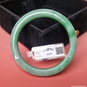 Könnte beinhalten: Ein glatter, kreisförmiger Armreif aus grüner Jadeit mit durchscheinender Optik. An dem Armreif ist ein kleines weißes rechteckiges Etikett befestigt, das den Text EGL, NATURAL JADEITE JADE, die Nummer 222373 und einen QR-Code enthält.