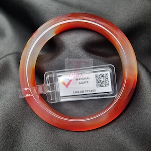 Puede incluir: Un brazalete de ágata translúcida, naranja y blanca. El brazalete es circular y tiene una superficie lisa y pulida. Una etiqueta de plástico transparente con las palabras "NATURAL AGATE" y "LULAB 575253" está adjunta.