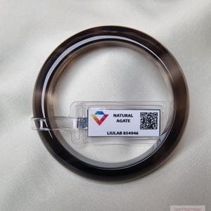Puede incluir: Un brazalete de ágata marrón oscuro translúcido. El brazalete tiene una superficie lisa y pulida y una forma circular. Una etiqueta con el texto "NATURAL AGATE" y "LIULAB 854946" está adjunta.
