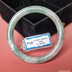 May include: A smooth, circular jadeite bangle featuring a translucent pale green and white mottled pattern. A small rectangular tag is attached to the bangle, displaying the text P-lab, NATURAL JADEITE, JADE, and N: 344109 in black ink.