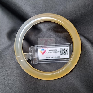 Può includere: Un braccialetto a cerchio giallo pallido e traslucido. Il braccialetto liscio e circolare è realizzato in calcedonio naturale. Un'etichetta trasparente con la scritta "NATURAL CHALCEDONY" e un codice QR è attaccata. Lo sfondo è un tessuto grigio scuro.