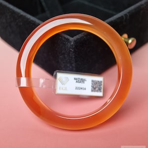 Puede incluir: Un brazalete rígido de ágata naranja, liso y translúcido. Una pequeña etiqueta rectangular blanca está adherida al brazalete, con el texto "NATURAL AGATE", "EGL", "222416" y un código QR. El brazalete tiene un acabado pulido y brillante.