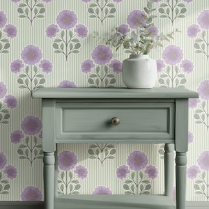 May include: A sage green wooden side table with a single drawer and a round bronze knob. On top of the table sits a white ceramic vase containing green leafy stems. The background features wallpaper with a vertical stripe and floral pattern.