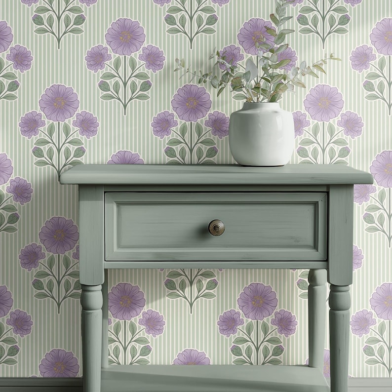 May include: A sage green wooden side table with a single drawer and a round bronze knob. On top of the table sits a white ceramic vase containing green leafy stems. The background features wallpaper with a vertical stripe and floral pattern.