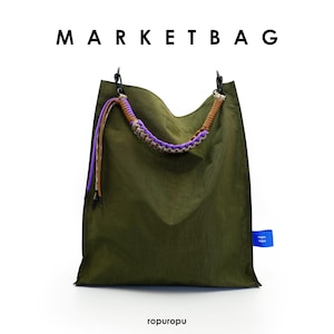 Op de afbeelding: Een olijfgroene markttas met de tekst MARKET BAG en ropuropu erop gedrukt. De tas heeft een gevlochten handvat in paars en beige, zwarte metalen clips en een klein blauw zijlabel met witte tekst. Het ontwerp is eenvoudig en functioneel.
