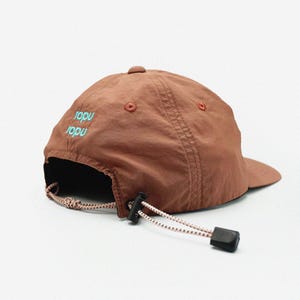 May include: A brown baseball cap with a curved peak. The cap has a small, round button on top and two small eyelets on the side. The text "Iopu" is embroidered in teal on the back. It features an adjustable strap with a black clasp.