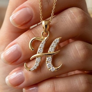 May include: A gold-toned necklace featuring a cursive letter H pendant. The pendant is embellished with small, clear, sparkling stones set along the surface of the letter. The necklace includes a delicate gold-toned chain with a standard link design.