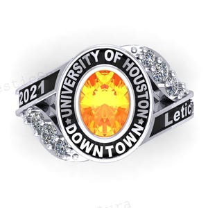 May include: A silver ring with an oval, yellow gemstone at the centre. The ring features the text "UNIVERSITY OF HOUSTON DOWNTOWN" and "2021" and "Letic" in black lettering. Small clear stones are set on either side of the central stone.