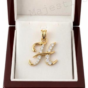 May include: A gold letter H pendant featuring a polished finish and a series of small, clear, round-cut stones embedded along the front surface. The pendant includes a gold bail at the top and is presented inside a dark wood jewellery box.