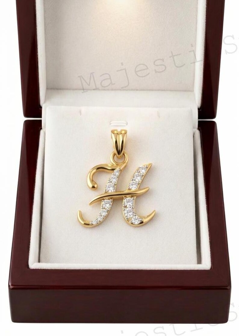 May include: A gold letter H pendant featuring a polished finish and a series of small, clear, round-cut stones embedded along the front surface. The pendant includes a gold bail at the top and is presented inside a dark wood jewellery box.