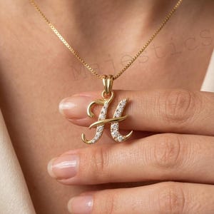 May include: A gold-toned necklace featuring a cursive letter H pendant. The pendant is embellished with small, clear, sparkling stones set along the front surface of the letter. The necklace includes a delicate gold-toned chain with a box link design.