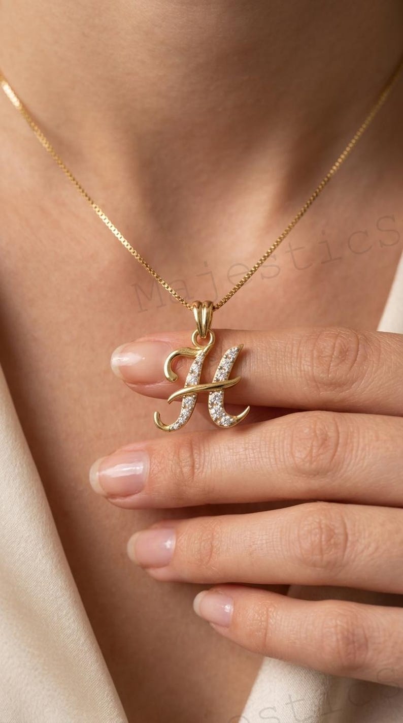 May include: A gold-toned necklace featuring a cursive letter H pendant. The pendant is embellished with small, clear, sparkling stones set along the front surface of the letter. The necklace includes a delicate gold-toned chain with a box link design.