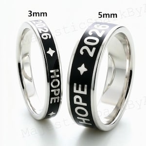 Puede incluir: Dos anillos plateados con bandas de esmalte negro. Los anillos muestran la palabra "HOPE" y el año "2026" en letras blancas, junto con un símbolo de estrella. Un anillo mide 3 mm de ancho, el otro 5 mm.