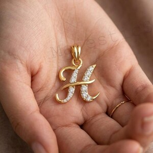May include: A gold pendant in the shape of the letter H, featuring a polished gold finish on the left side and a series of small, clear, sparkling stones embedded along the right side of the letter, complete with a gold bail at the top.