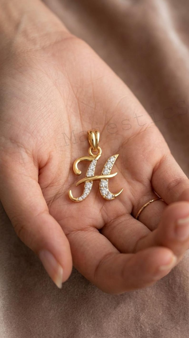 May include: A gold pendant in the shape of the letter H, featuring a polished gold finish on the left side and a series of small, clear, sparkling stones embedded along the right side of the letter, complete with a gold bail at the top.
