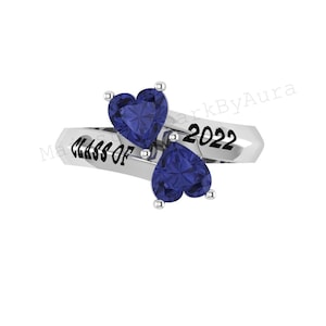 May include: A silver ring with two heart-shaped, dark blue gemstones. The ring is inscribed with "CLASS OF 2022". The gemstones are set on a diagonal, creating a unique design. A graduation-themed jewelry item.