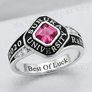 May include: A silver ring with a square pink gemstone at the center, surrounded by a black border with the words "SURUKA UNIVERSITY" and "2020". The ring also features small clear stones and the inscription "Best Of Luck".
