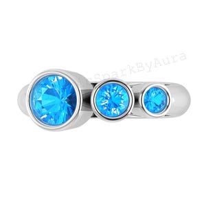 Puede incluir: Un anillo de plata con tres piedras preciosas redondas de color azul brillante. Las piedras están engastadas en un bisel y el anillo tiene un acabado liso y pulido. El anillo se muestra sobre un fondo blanco.