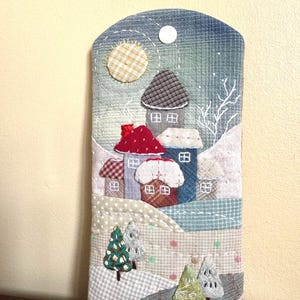 May include: A rectangular fabric case featuring a quilted winter village scene with houses in red, blue, and brown, a yellow sun, and green and grey trees. The design includes white stitching for snow and a white snap button at the top center.