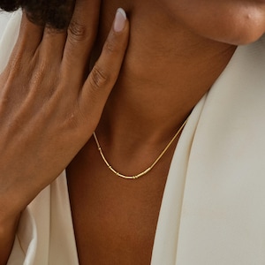 Peut inclure: Un délicat collier en or au design minimaliste. Le collier est composé d'une fine chaîne avec de petites perles cylindriques et sphériques espacées régulièrement. Le bijou est porté sur une veste blanche.