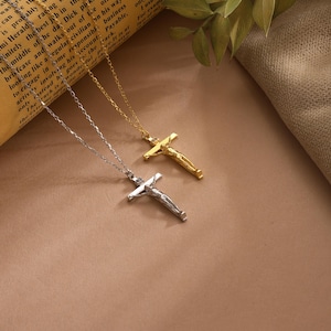 May include: Two crucifix necklaces, one silver and one gold, displayed on a brown surface. The pendants depict a figure on a cross. The necklaces are on a chain. The background includes a rolled-up paper with text and some greenery.